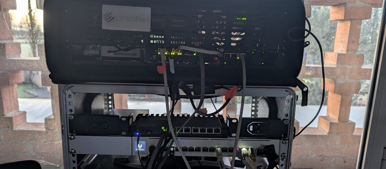 Il kit LinkoNet aperto e collegato davanti a una finestra con muratura in mattoni: apparati con LED accesi, cavi di rete e switch montati nel rack